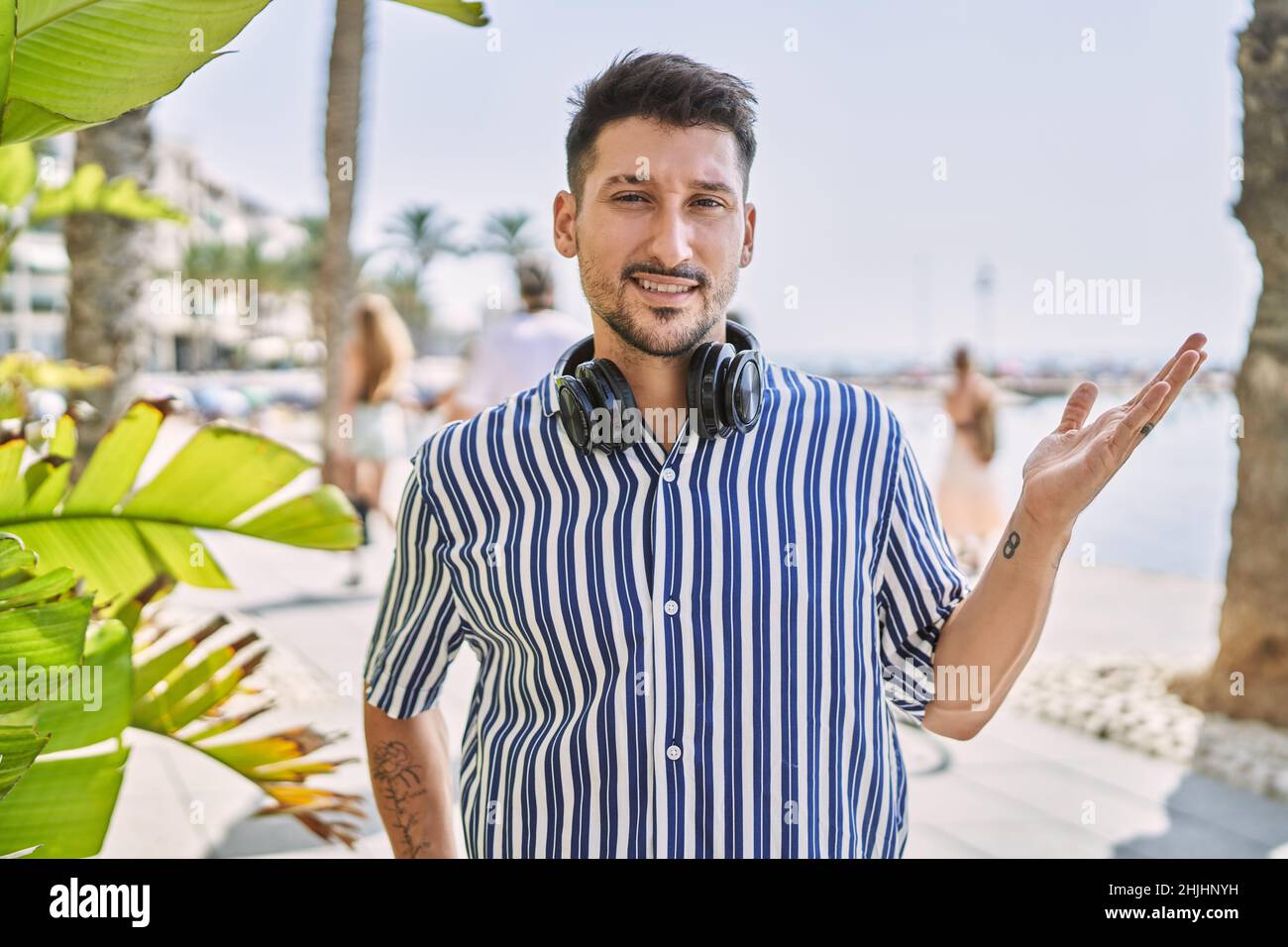 Junger, hübscher Mann, der im Freien mit Kopfhörern Musik hört, lächelt fröhlich und zeigt mit der Handfläche, die auf die Kamera schaut. Stockfoto