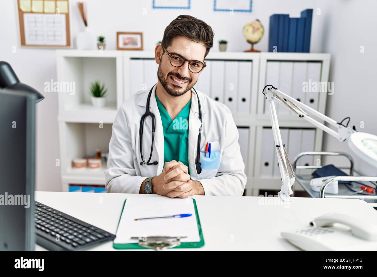 Junger Mann mit Bart in der Klinik mit Arztuniform und Stethoskop, die Hände zusammen und die gekreuzten Daumen lächeln entspannt und fröhlich. Erfolg Stockfoto