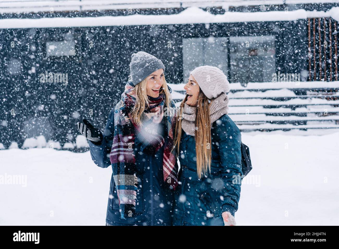 Zwei junge Frauen in warmer Winterkleidung stehen unter dem Schneefall und lachen fröhlich. Stockfoto