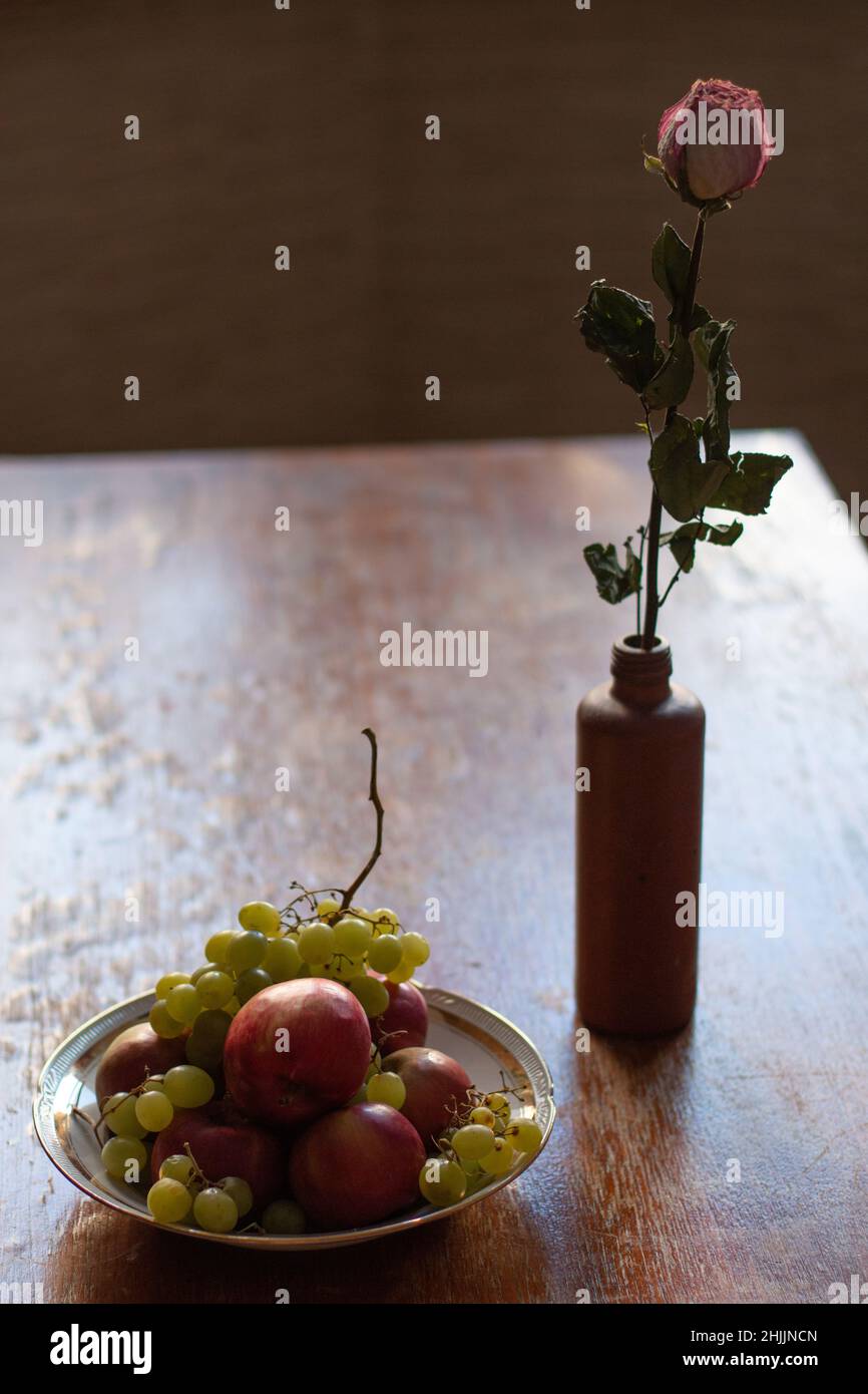 Trockene Rosenblüte in einem Tongefäß, neben einer Schale mit Früchten, Trauben und Äpfeln, auf einem roten Holztisch, Stillleben, vertikales Foto Stockfoto