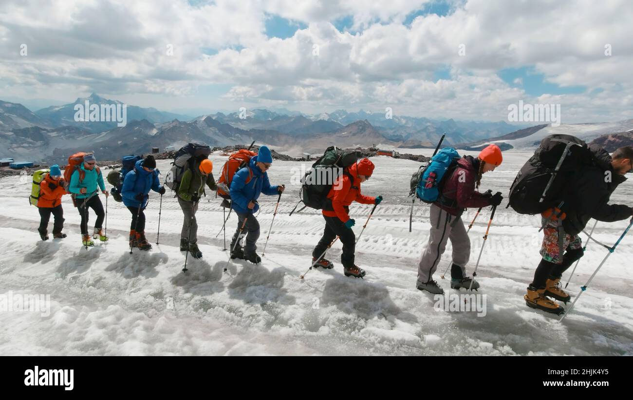 Kletterer besteigen den Berg vor dem Hintergrund der Berglandschaft. Clip. Eine Gruppe von Kletterern läuft an sonnigen Tagen mit Stöcken am Hang des verschneiten Berges. Stockfoto