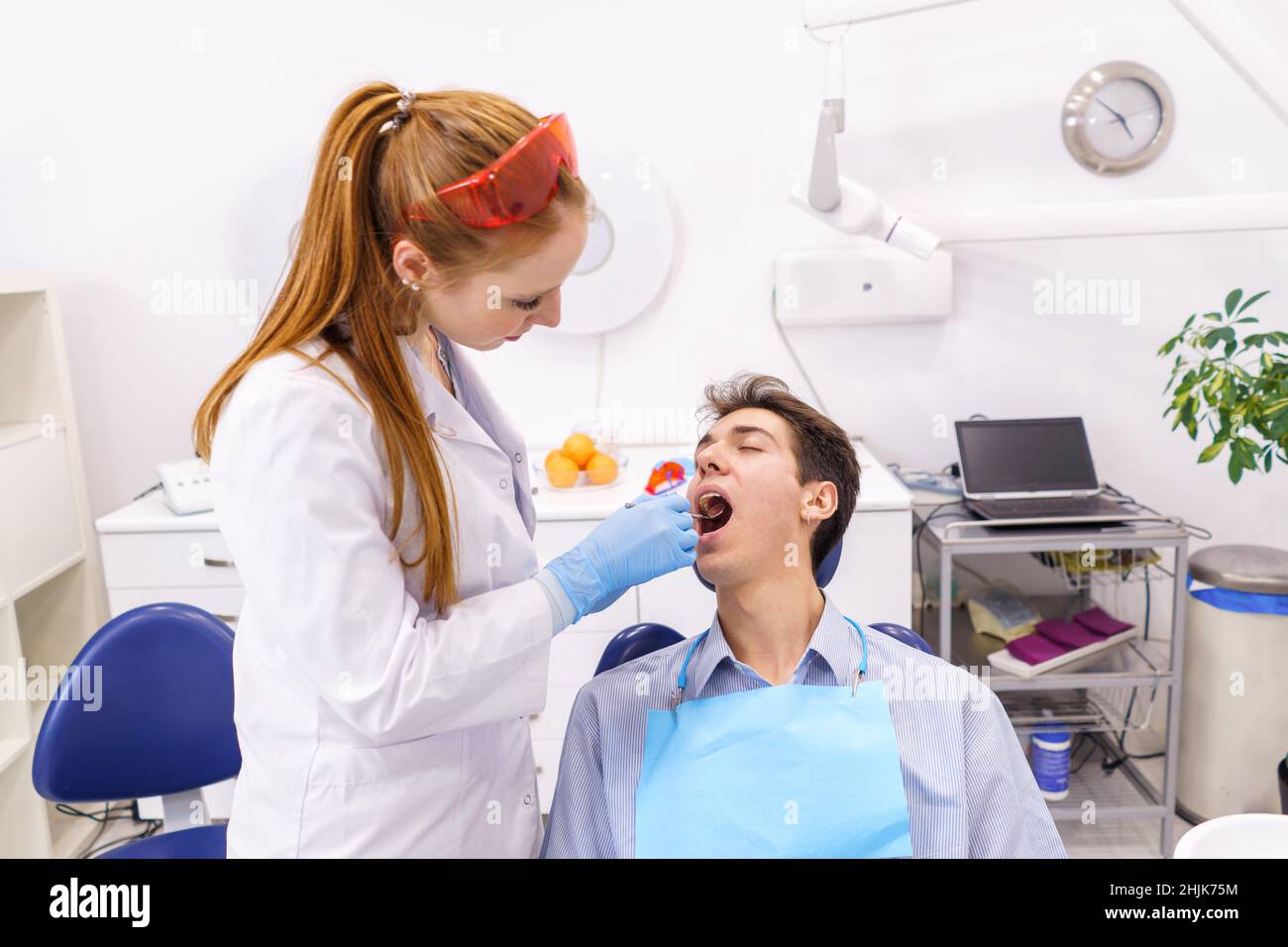 Ingwer Frau in weißem Gewand und Latexhandschuhe mit Spiegel, um die Zähne des Mannes zu untersuchen, während sie in einem modernen Büro der Zahnklinik arbeitet Stockfoto