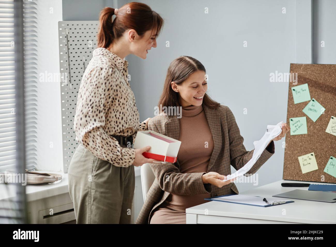 Glückliche, schwanger Frau, die an ihrem Arbeitsplatz sitzt und die Kleidung auspackt, die ihr Kollege für ihr Baby gibt Stockfoto