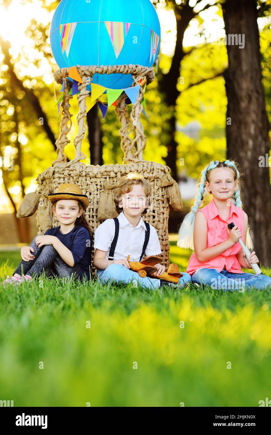 Drei kleine Vorschulkinder sitzen auf dem Gras vor dem Hintergrund eines Korbes aus blauen Ballons und Sonnenlicht. Stockfoto