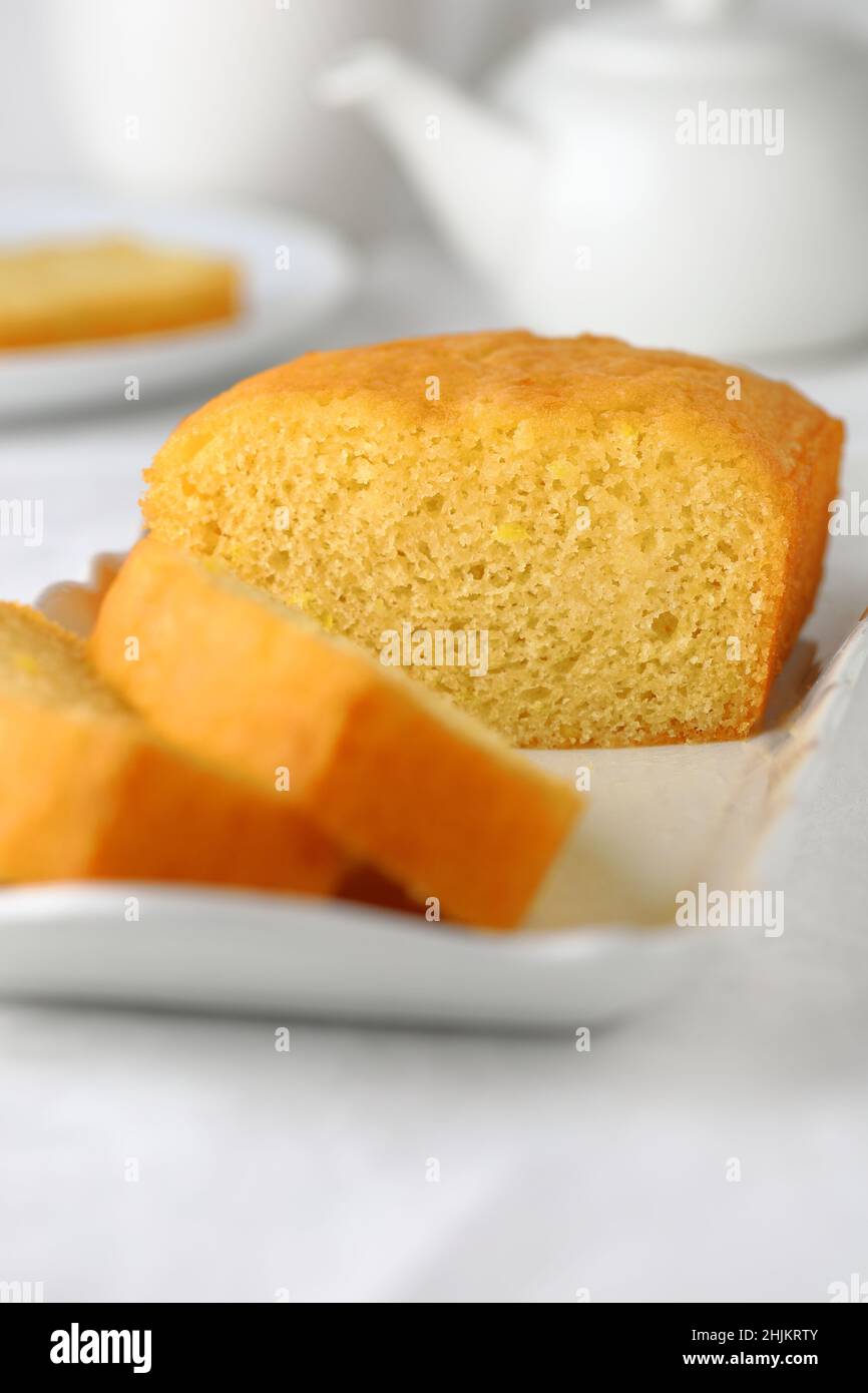 Selbstgebackener Kuchen für den Nachmittagstee, in Scheiben geschnitten auf einem weißen rechteckigen Teller mit weißer Teekane und Tasse im Hintergrund Stockfoto