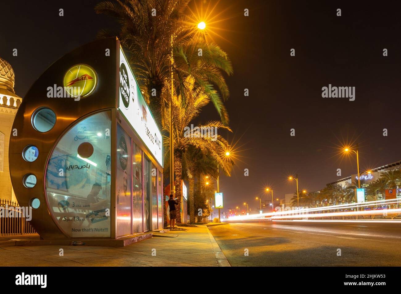 Dubai, VAE, 27.09.21. Klimatisierte RTA-Bushaltestelle in Dubai bei Nacht auf einer belebten Straße, mit Ampelstreifen und Palmen, Langzeitbelichtung Foto. Stockfoto