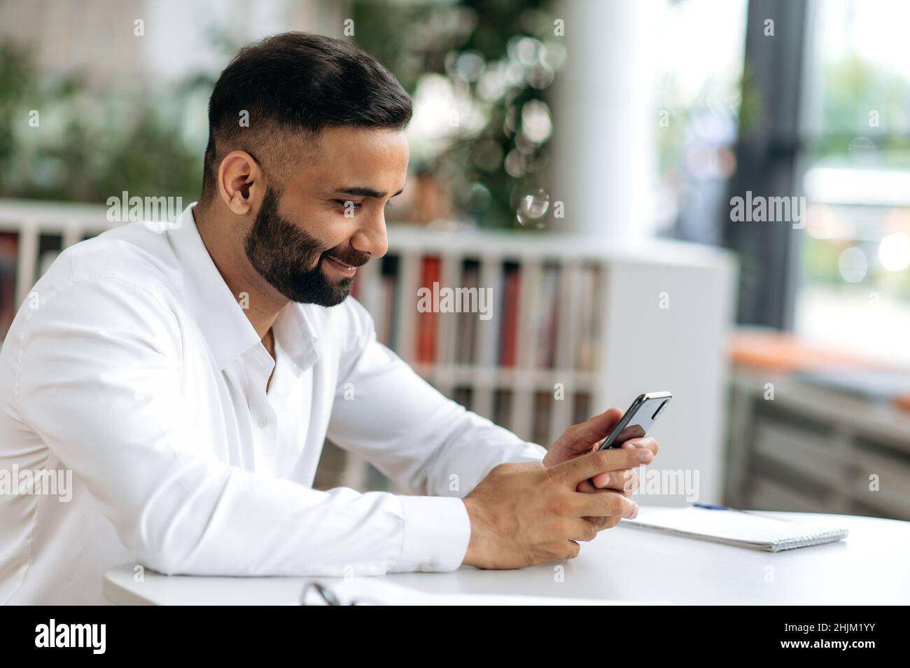 SMS-Bearbeitung. Ein sympathischer, erfolgreicher Inder, ein Händler, ein Top-Manager, sitzt an seinem Schreibtisch, führt während der Arbeitszeit Arbeitskorrespondenz auf einem Mobiltelefon durch und lächelt freundlich Stockfoto