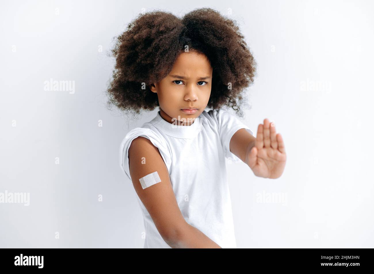 Trauriges afroamerikanisches, niedliches kleines Mädchen, das nach der Impfung ein Pfand auf der Schulter hatte und eine STOP-Geste gegen die Impfung von Kindern zeigte, steht auf einem isolierten weißen Hintergrund Stockfoto