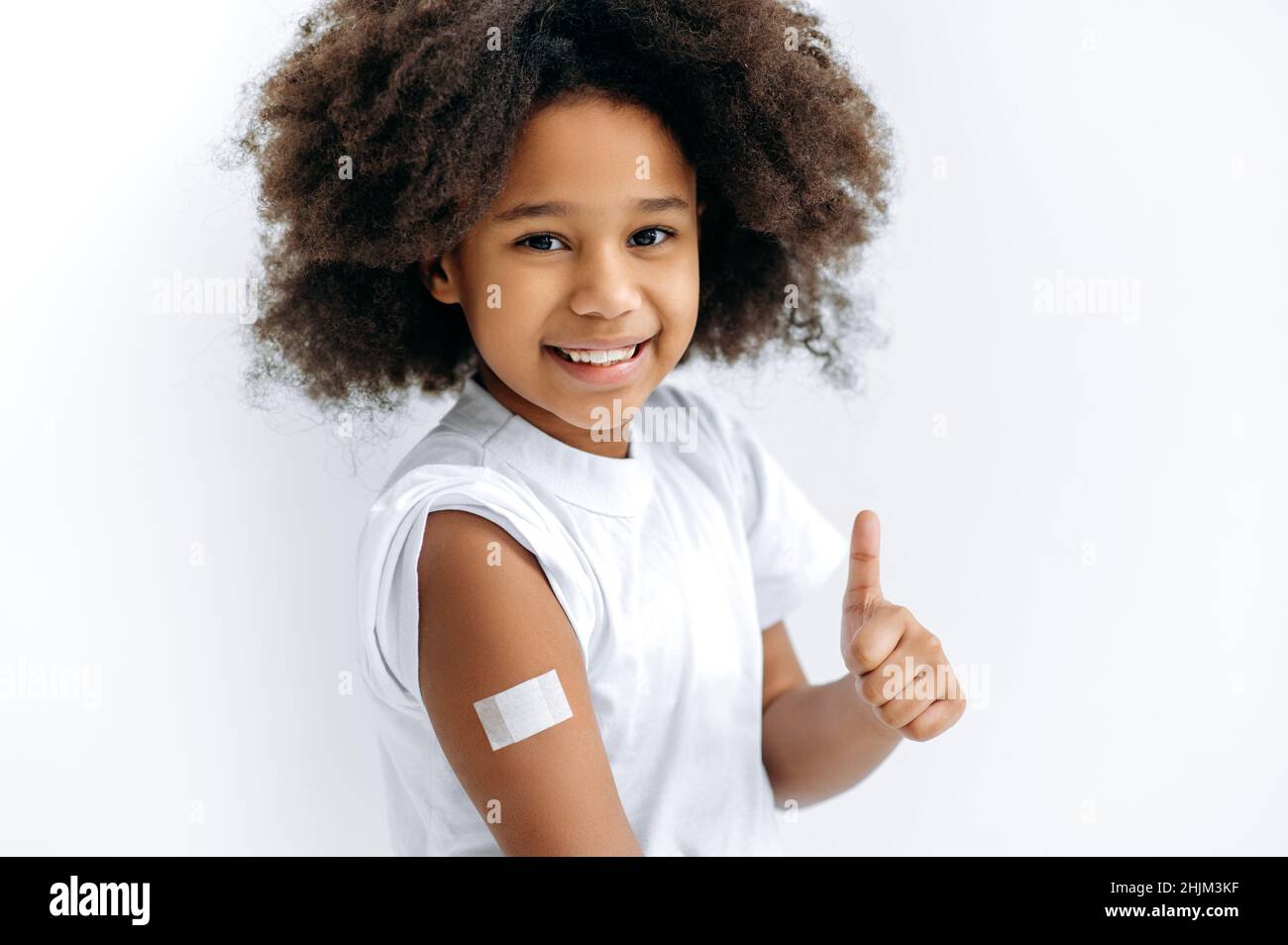 Glückliches afroamerikanisches nettes kleines Mädchen, Vorschulkinder, mit einem Pfand auf ihrer Schulter nach der Impfung, für Immunität gegen covid 19 und andere Krankheiten, stehen auf isoliertem weißen Hintergrund, Daumen-up Stockfoto