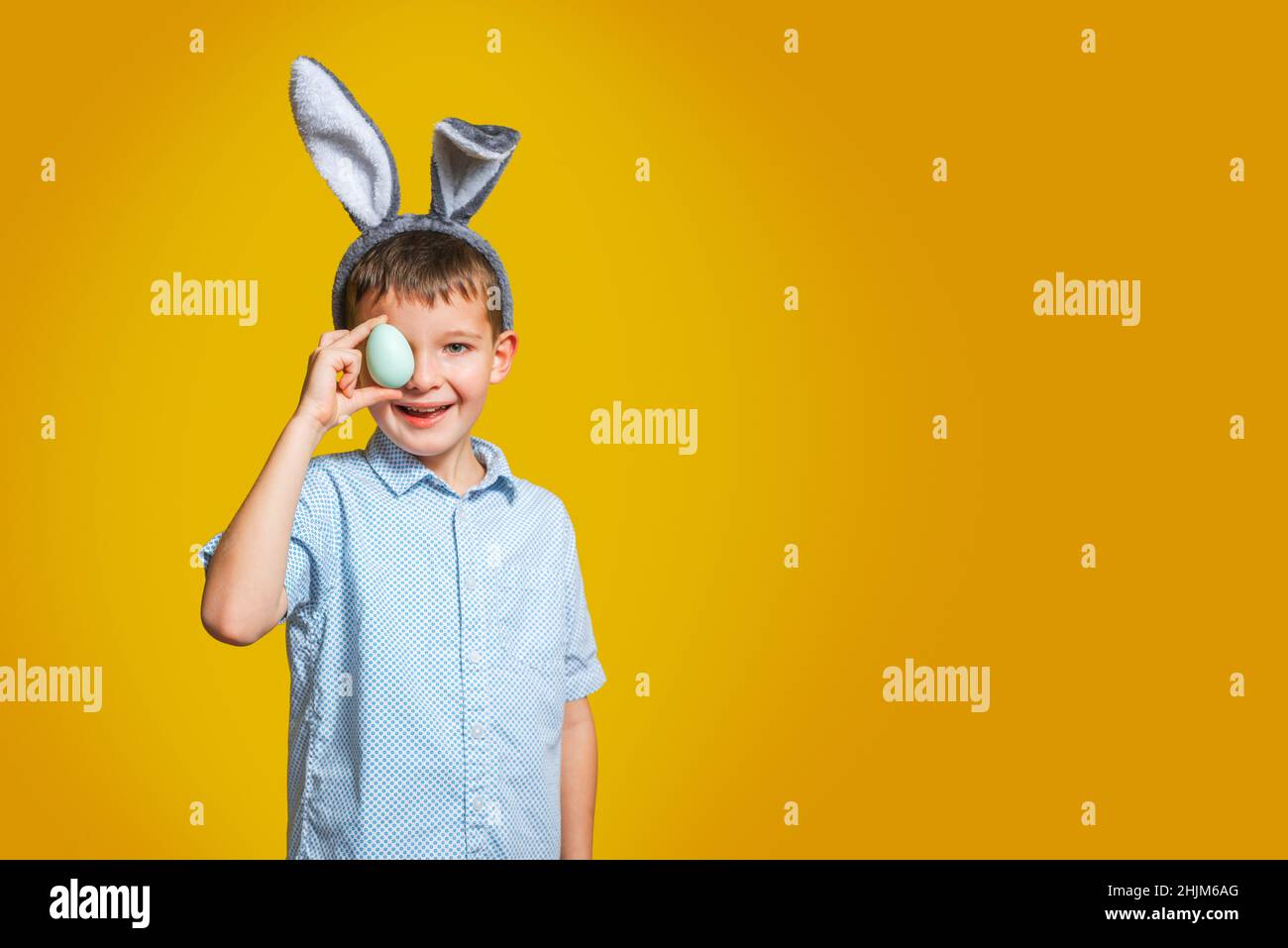 Glücklicher Junge mit Hasenohren bedeckt sein Auge mit Osterei auf gelbem Hintergrund. Ein Kind mit Osterei auf gelbem Hintergrund. Stockfoto