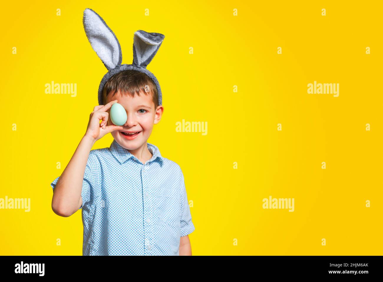 Glücklicher Junge mit Hasenohren bedeckt sein Auge mit Osterei auf gelbem Hintergrund. Ein Kind mit Osterei auf gelbem Hintergrund. Stockfoto