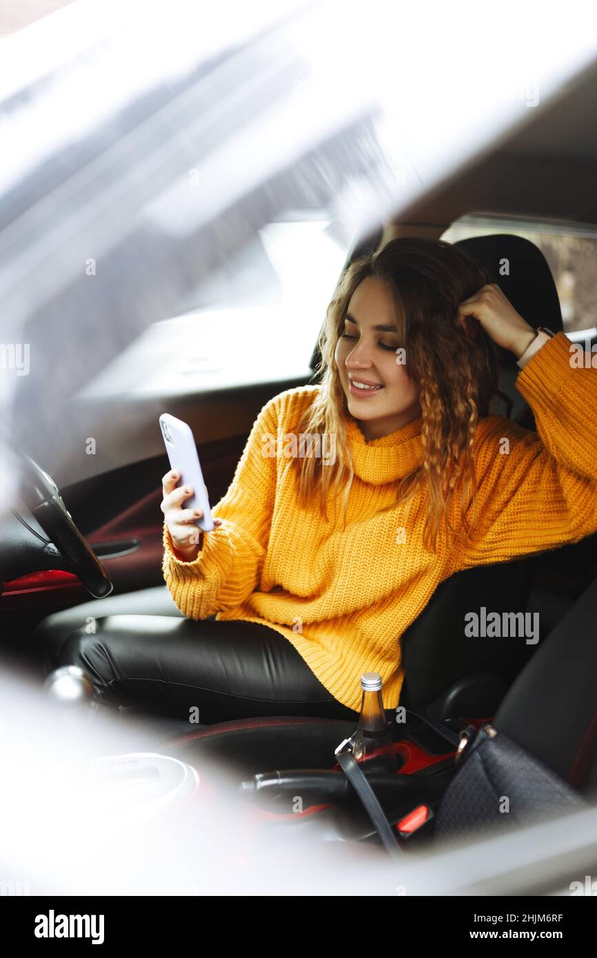 Porträt einer jungen, lässigen Frau in orangefarbenem Pullover, SMS auf ihrem Smartphone oder Videoanruf, während der Fahrt oder der Verkehr ein Auto während der Mittagspause anhalten. Stockfoto