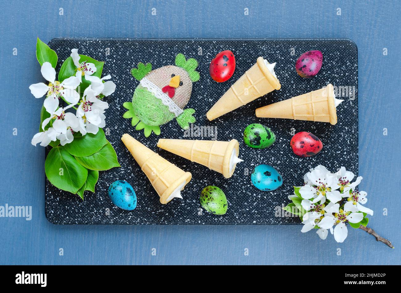 Ostern buntes Stillleben auf einem Schneidebrett mit bunten Wachteleiern, Dessert in Waffelkegel und einem Spielzeug-Huhn auf einem Hintergrund von Fruchtinflorescen Stockfoto