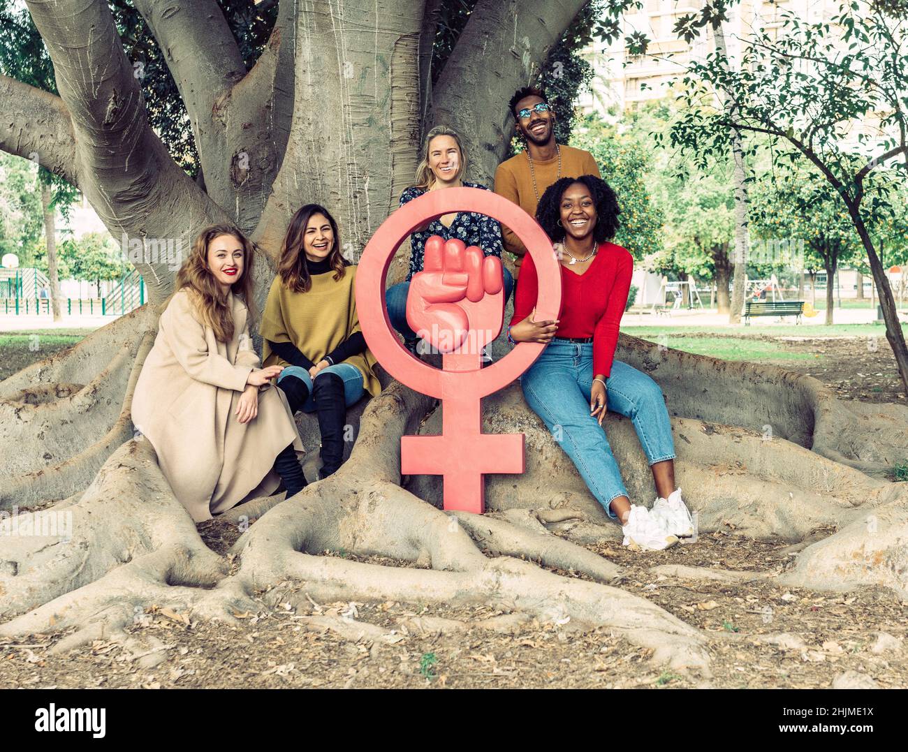 5 Menschen lächeln neben einem Ficus macrophylla um eine große Ikone, die Feminismus symbolisiert. Stockfoto