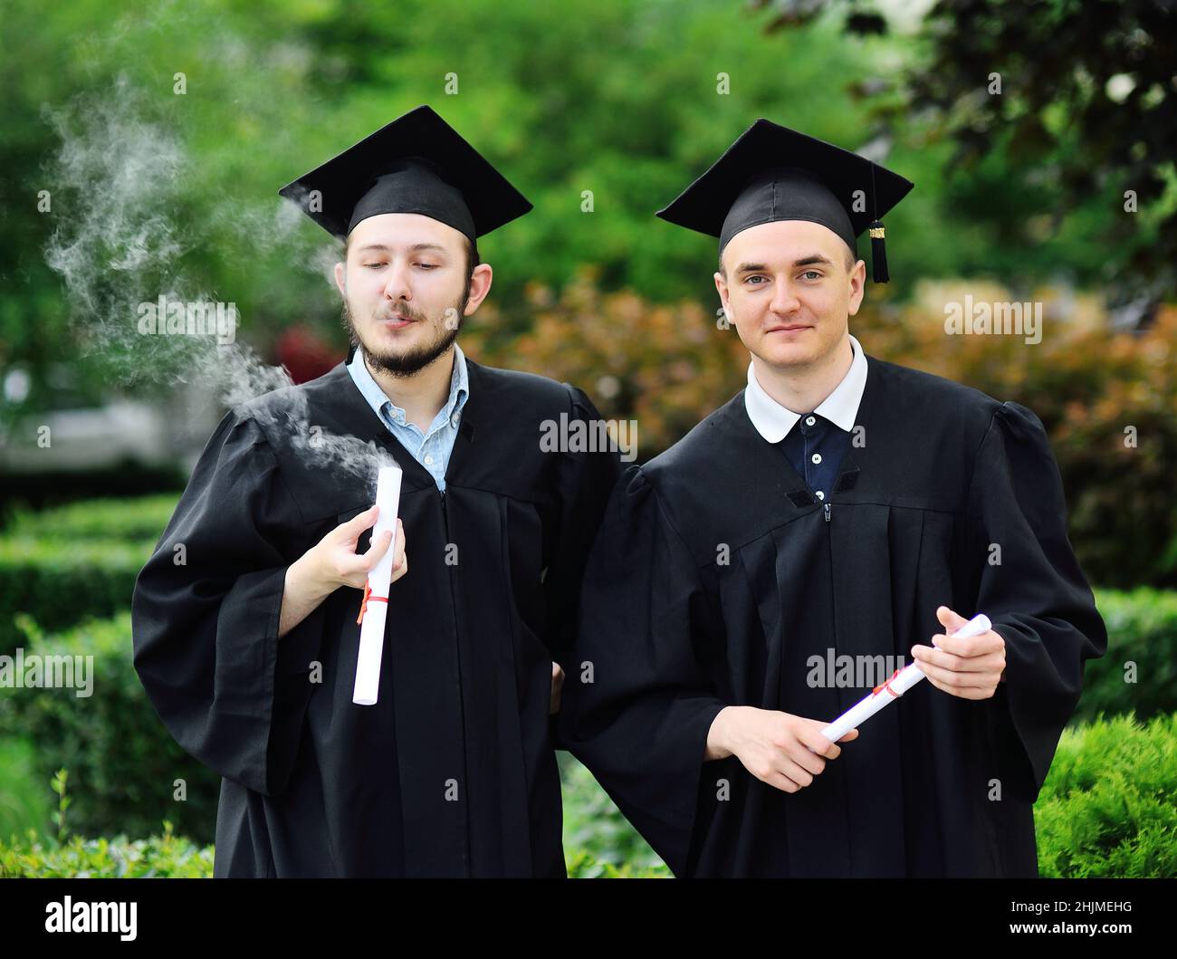 Ein Doktorand in einem schwarzen Gewand und einem quadratischen Hut raucht sein Diplom oder Zertifikat. Marihuana, Cannabis und Studentenleben. Stockfoto