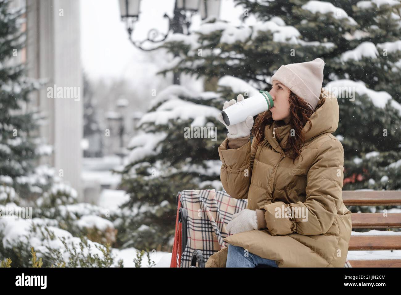 Fröhlich brünette Frau trinken aus Kolben im Freien in der Stadt im Winter. Aufwärmen, Momente genießen, Bremsen über schneebedeckten Bäumen Stockfoto