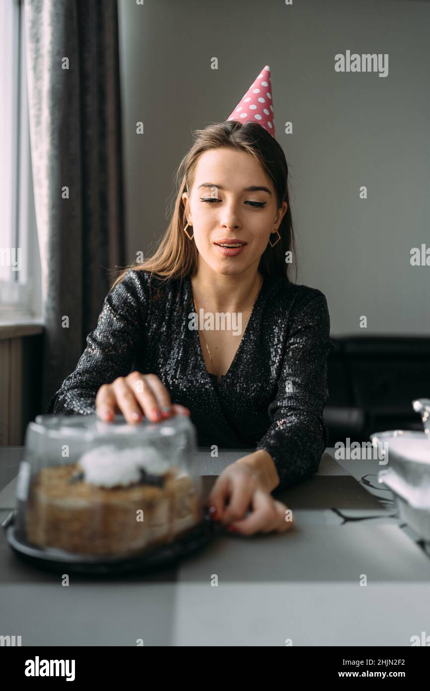 Geburtstag zu Hause. Junge glückliche Frau öffnet eine Schachtel mit einem Kuchen. Ein Mädchen in einer festlichen Mütze und kleiden sich am Tisch. Wandhintergrund. Geburtstagsfeier Stockfoto