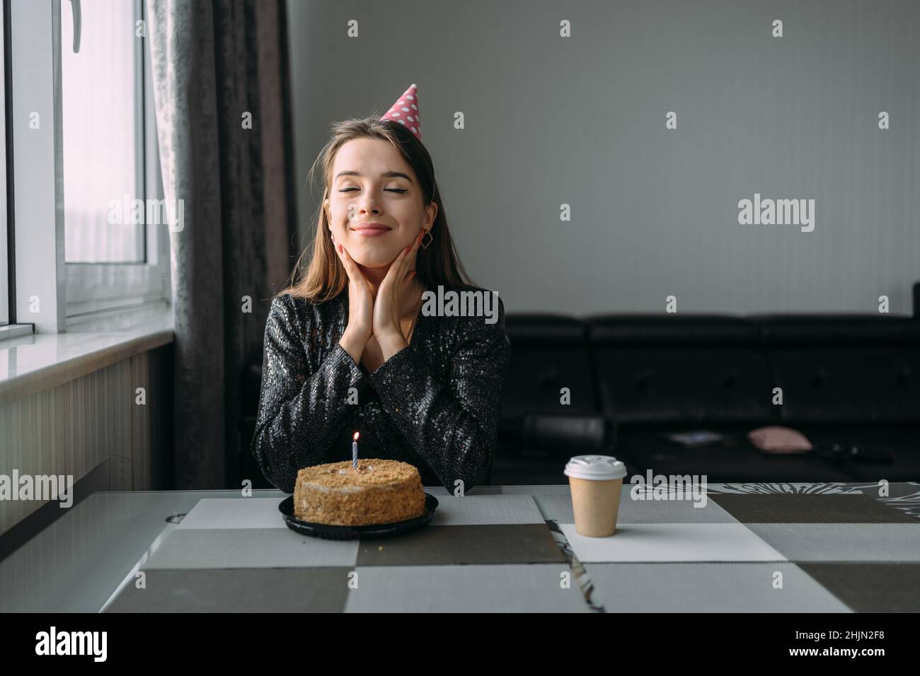 Geburtstag zu Hause. Junge glückliche Frau mit einem Kuchen am Tisch. Das Mädchen in festlicher Mütze und Kleid ist allein. Wandhintergrund. Geburtstagsfeier Stockfoto