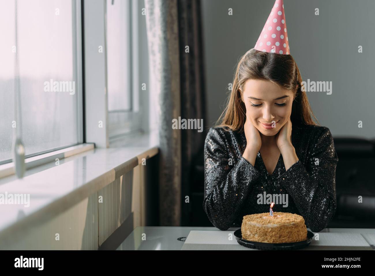 Porträt einer traurigen jungen Frau, die an ihrem Geburtstag am Tisch sitzt, eines einsamen Urlaubs zu Hause, eines Mädchens in einer Mütze, das in Quarantäne ein Jubiläum feiert Stockfoto