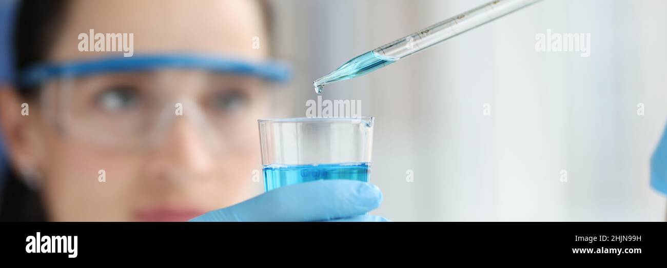 Weibliche Wissenschaftlerin tropft und pipettiert in einen Kolben aus blauer Flüssigkeit Stockfoto