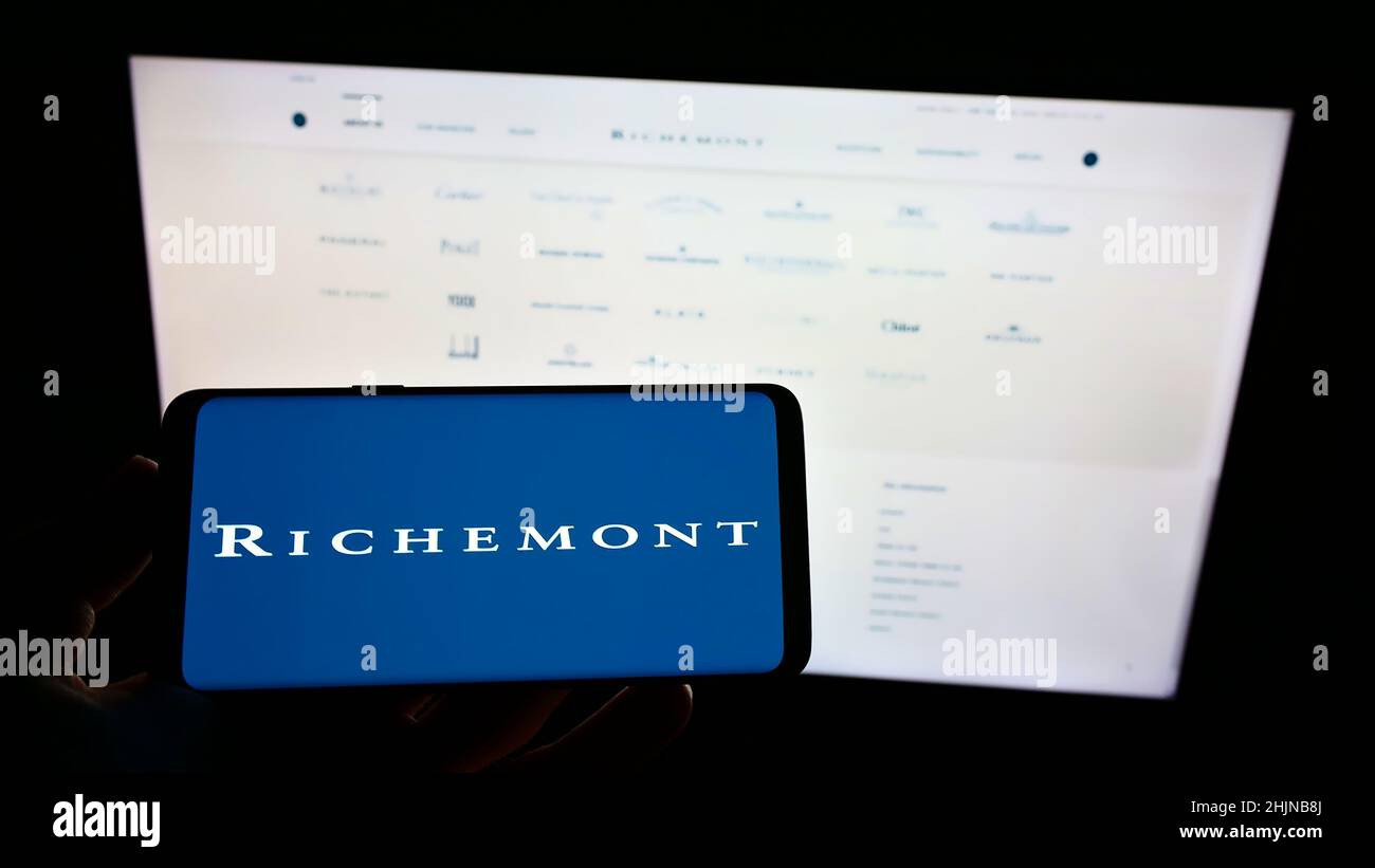 Person, die Mobiltelefon mit Logo der Schweizer Compagnie Financiere Richemont S.A. auf dem Bildschirm vor der Webseite hält. Konzentrieren Sie sich auf die Telefonanzeige. Stockfoto