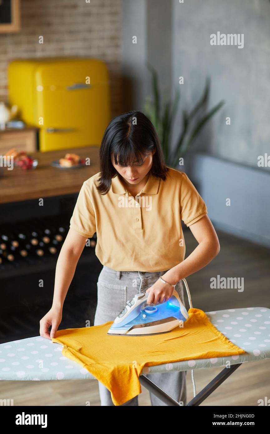 Vertikales Porträt einer jungen asiatischen Frau, die zu Hause Kleidung bügelt und Hausaufgaben macht Stockfoto