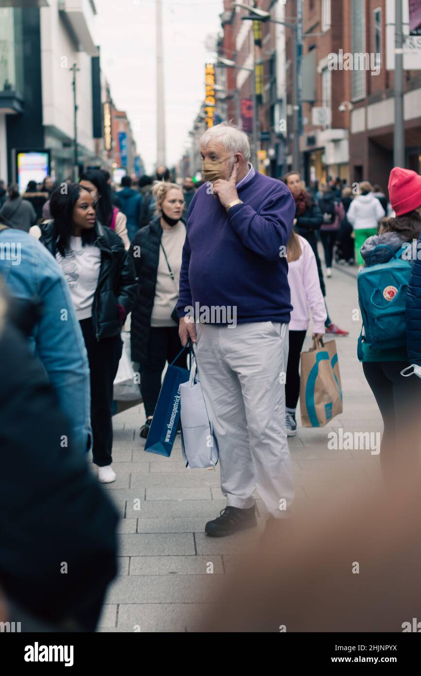 Große alte Männer mit Gesichtsmaske zwischen Menschenmengen an der Henry Street mit Einkaufstasche in der Hand, verschwommene Straßenfotografie im Hintergrund, Dublin, Irland Stockfoto