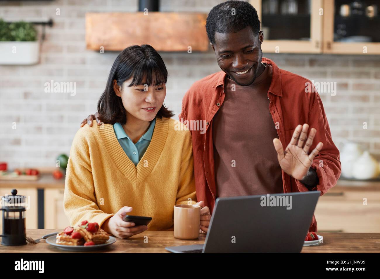 Waist-up-Porträt eines glücklichen Mixed-Race-Paares, das im Videochat in der gemütlichen, farbenfrohen Küche mit der Kamera winkt Stockfoto