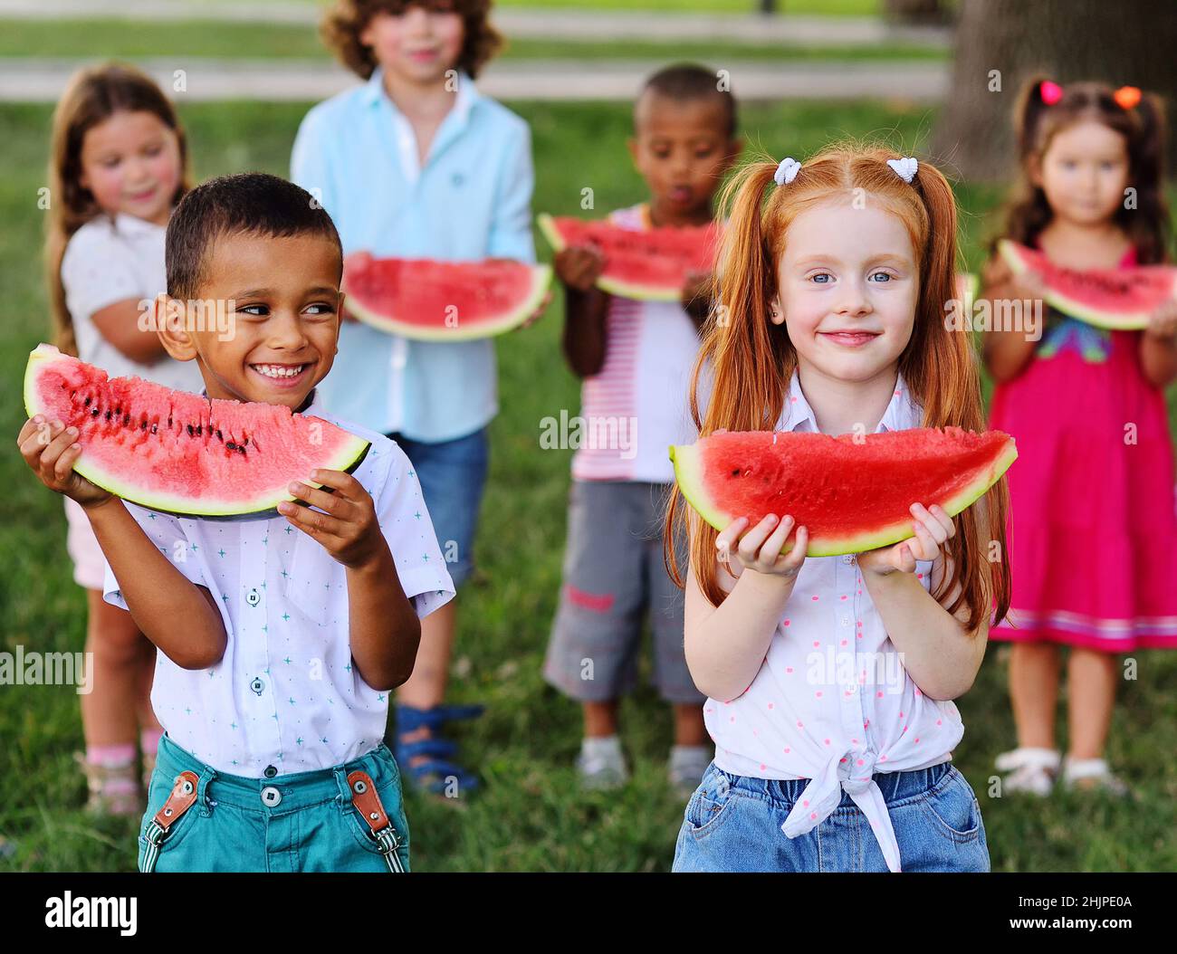 Eine Gruppe von glücklichen Vorschulkindern verschiedener ethnischer Gruppen hält reife Wassermelonen in Scheiben und lächelt im Hintergrund des Parks auf einem Sonnenbad Stockfoto