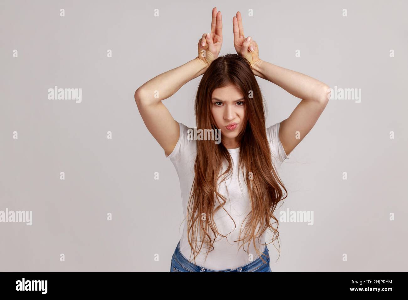 Porträt einer aggressiven, tyrannisierten Frau mit dunklen Haaren, die Stierhörner über den Kopf zeigt, wie vor dem Angriff Stirnrunzeln zeigt und weißes T-Shirt trägt. Innenaufnahme des Studios isoliert auf grauem Hintergrund. Stockfoto