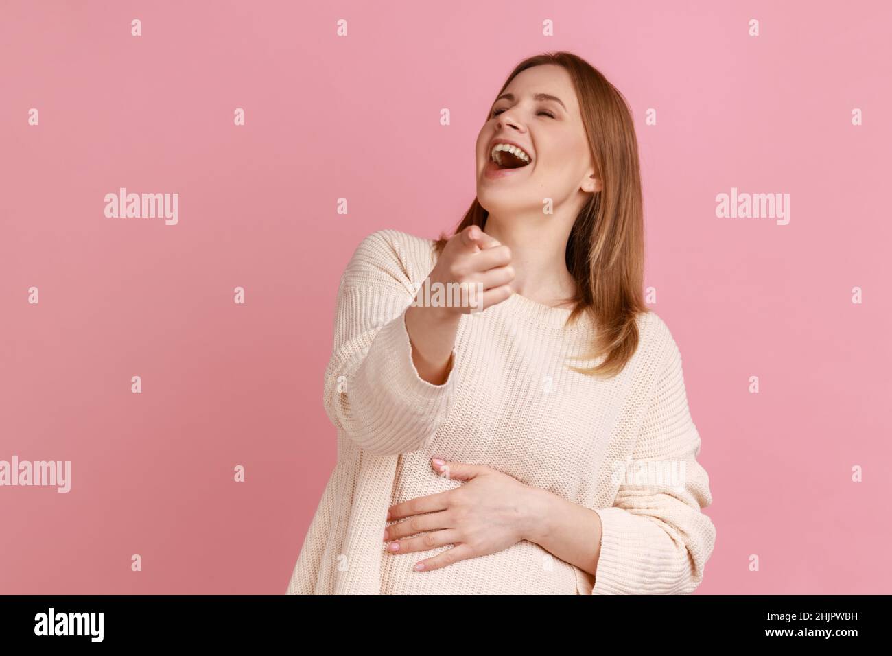 Porträt einer optimistischen blonden Frau, die laut mit dem Zeigefinger lacht, sich über Sie lustig macht und die Kamera anschaut, Witz, weißen Pullover trägt. Innenaufnahme des Studios isoliert auf rosa Hintergrund. Stockfoto