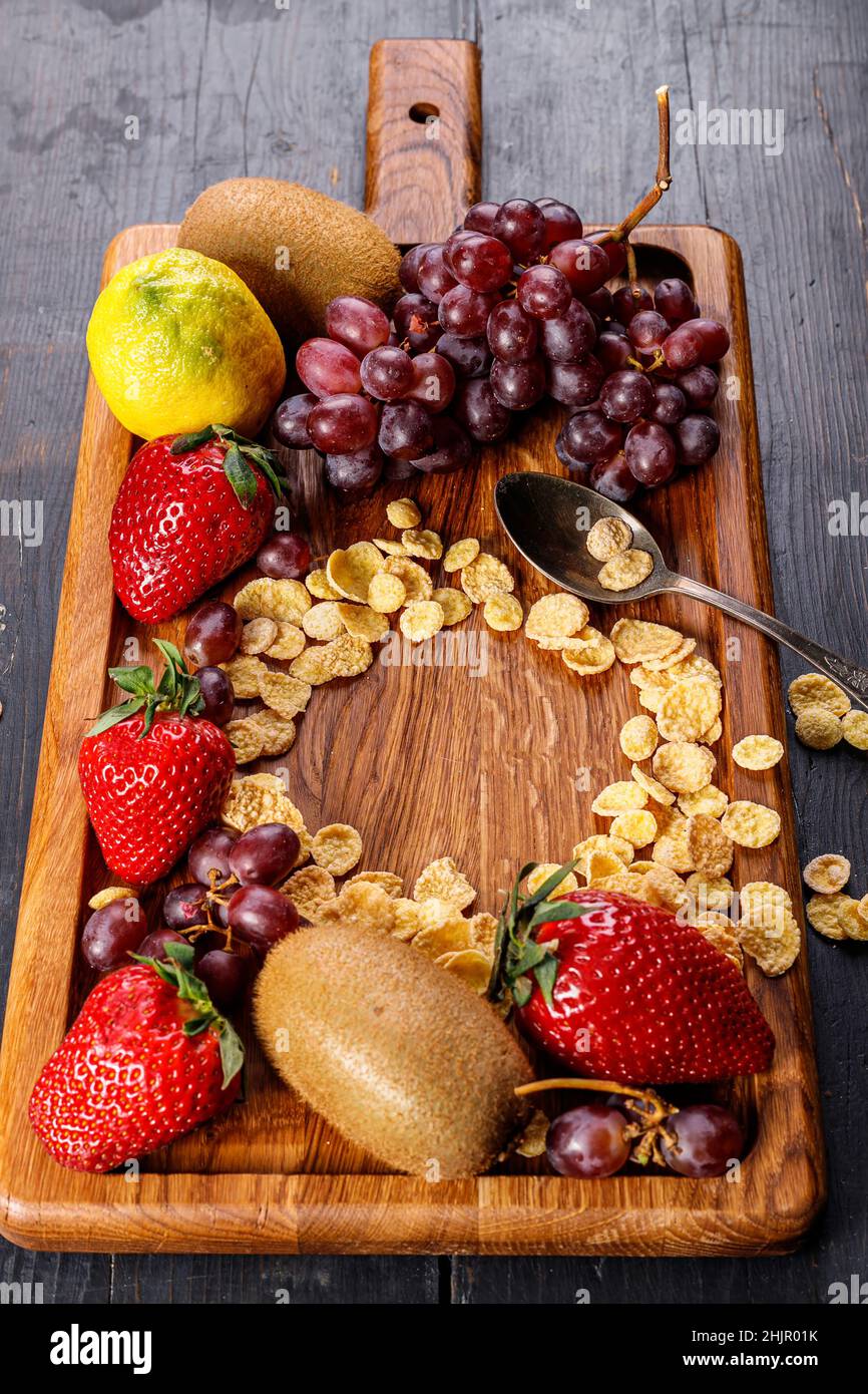 Cornflakes. Verschiedene saftige reife Früchte. Erdbeere, Kiwi, Trauben. Köstliche tropische Früchte auf einem hölzernen Hintergrund. Gesundes Frühstück. Vertikale Aufnahme. Stockfoto