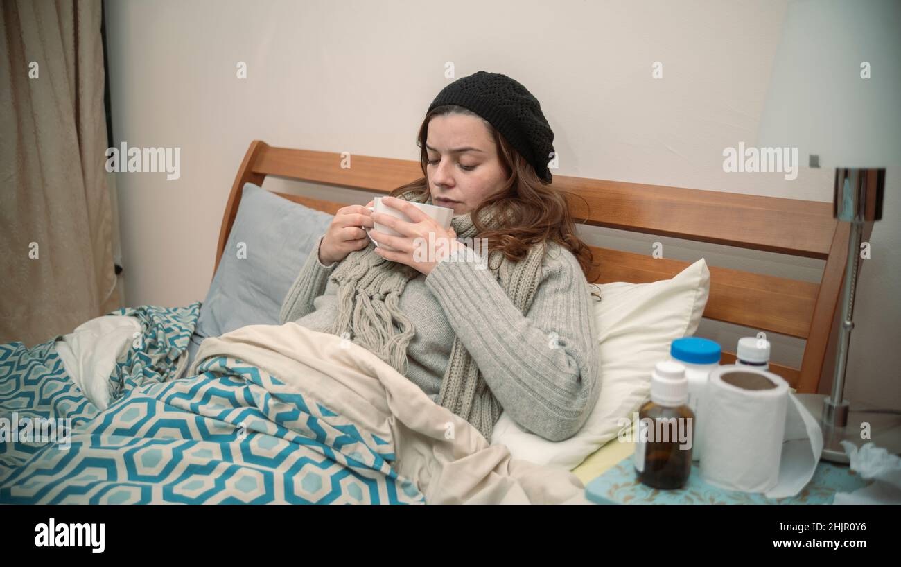 Hispanische Frau, die allein in ihrem gut gekleideten Bett neben einem Tisch voller Medikamente liegt, an der Grippe erkrankt ist und eine Tasse Tee trinkt Stockfoto