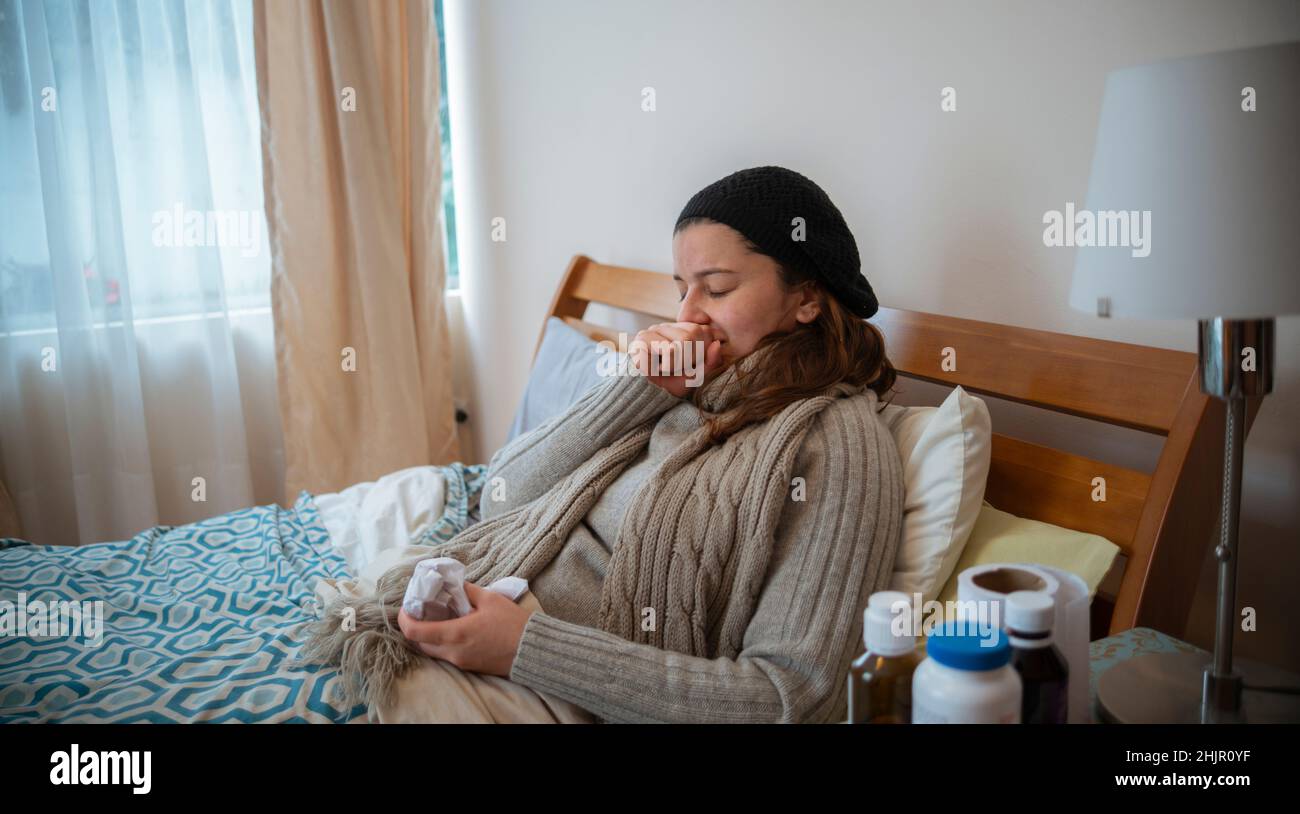 Hispanische Frau, die gut eingewickelt in ihrem Bett lag, an Grippe erkrankt, ihren Mund mit der Hand bedeckte, während sie niesen Stockfoto