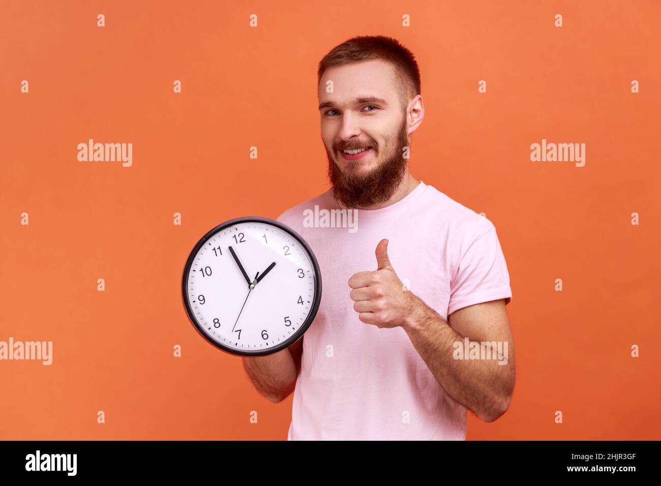 Porträt eines bärtigen Mannes, der die Daumen nach oben zeigt und eine große Wanduhr hält, mit einem Lächeln auf die Kamera blickt, Zeitmanagement, pinkes T-Shirt trägt. Innenaufnahme des Studios isoliert auf orangefarbenem Hintergrund. Stockfoto