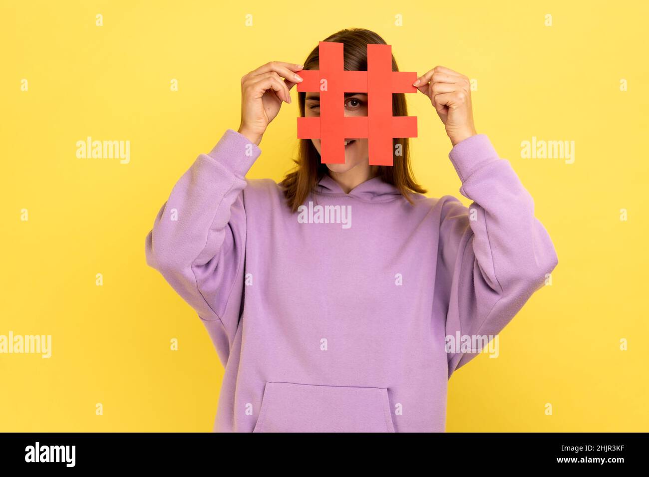 Porträt einer Frau, die Gesicht mit Social-Media-Hashtag-Symbol bedeckt, empfehlen, trendige Inhalte zu folgen, beliebten Blog, tragen lila Hoodie. Innenaufnahme des Studios isoliert auf gelbem Hintergrund. Stockfoto