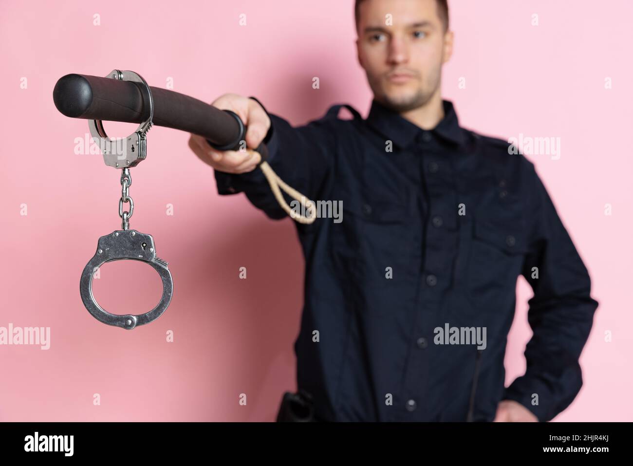 Ein gutaussehender Polizist in schwarzer Uniform mit Keule und Handschellen isoliert auf rosa Hintergrund. Stockfoto