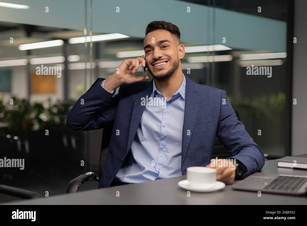 Fröhlicher arabischer Unternehmer sitzt am Arbeitstisch und telefoniert Stockfoto