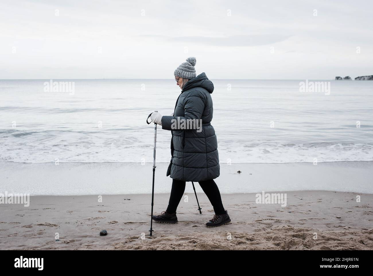 Frau in den Siebzigern, die im Winter mit Stöcken am Strand entlang läuft Stockfoto