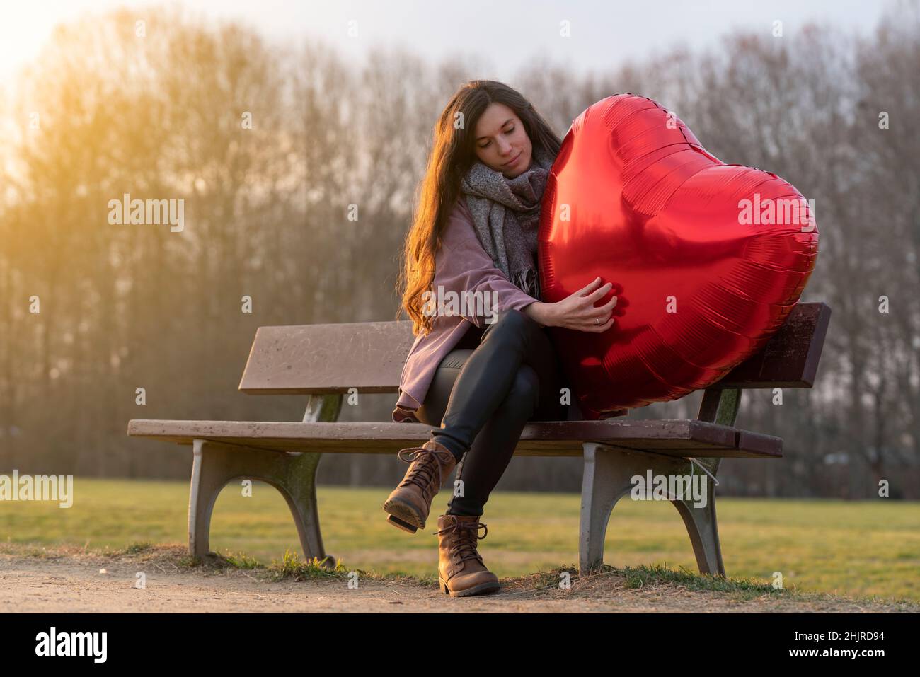 Traurige junge Frau, die einen herzförmigen Ballon umarmt und auf einer Bank im Park sitzt Stockfoto