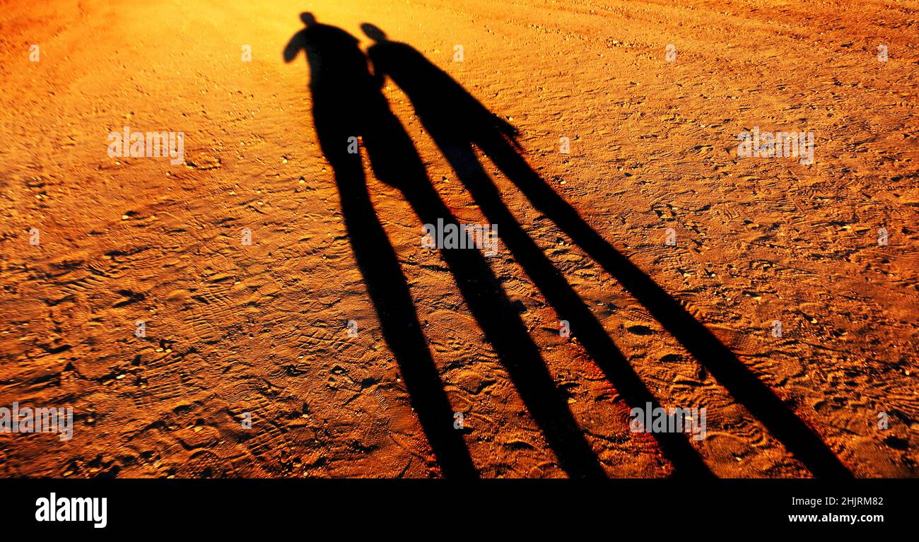 Lange Schatten von zwei Menschen, die sich die Hände halten, die zusammen eine unbefestigte Straße entlang gehen Stockfoto