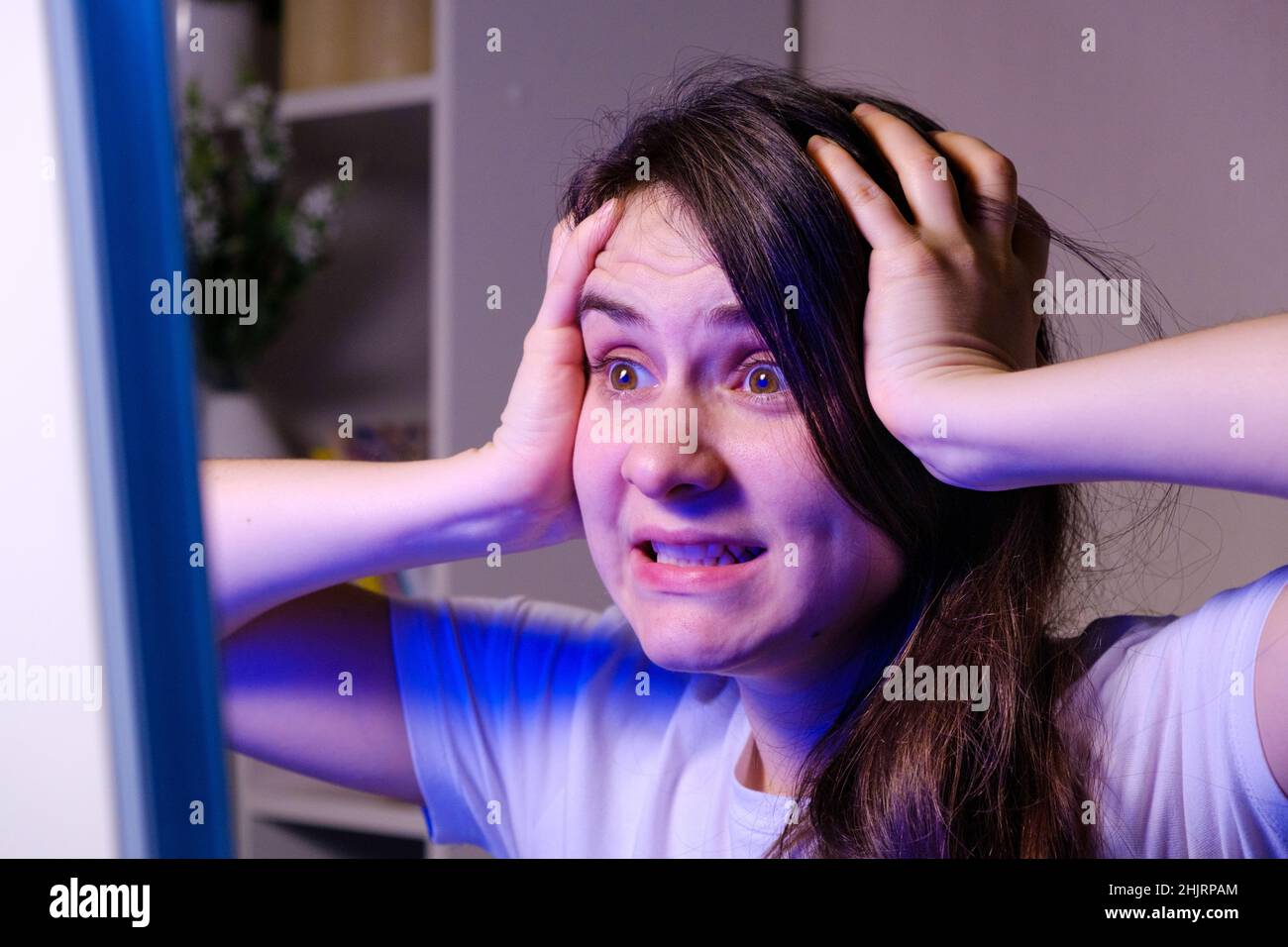 Eine Frau hält ihren Kopf mit ihren Händen und schaut auf den Fernseher oder den Computerbildschirm, ist sehr nervös. Stockfoto