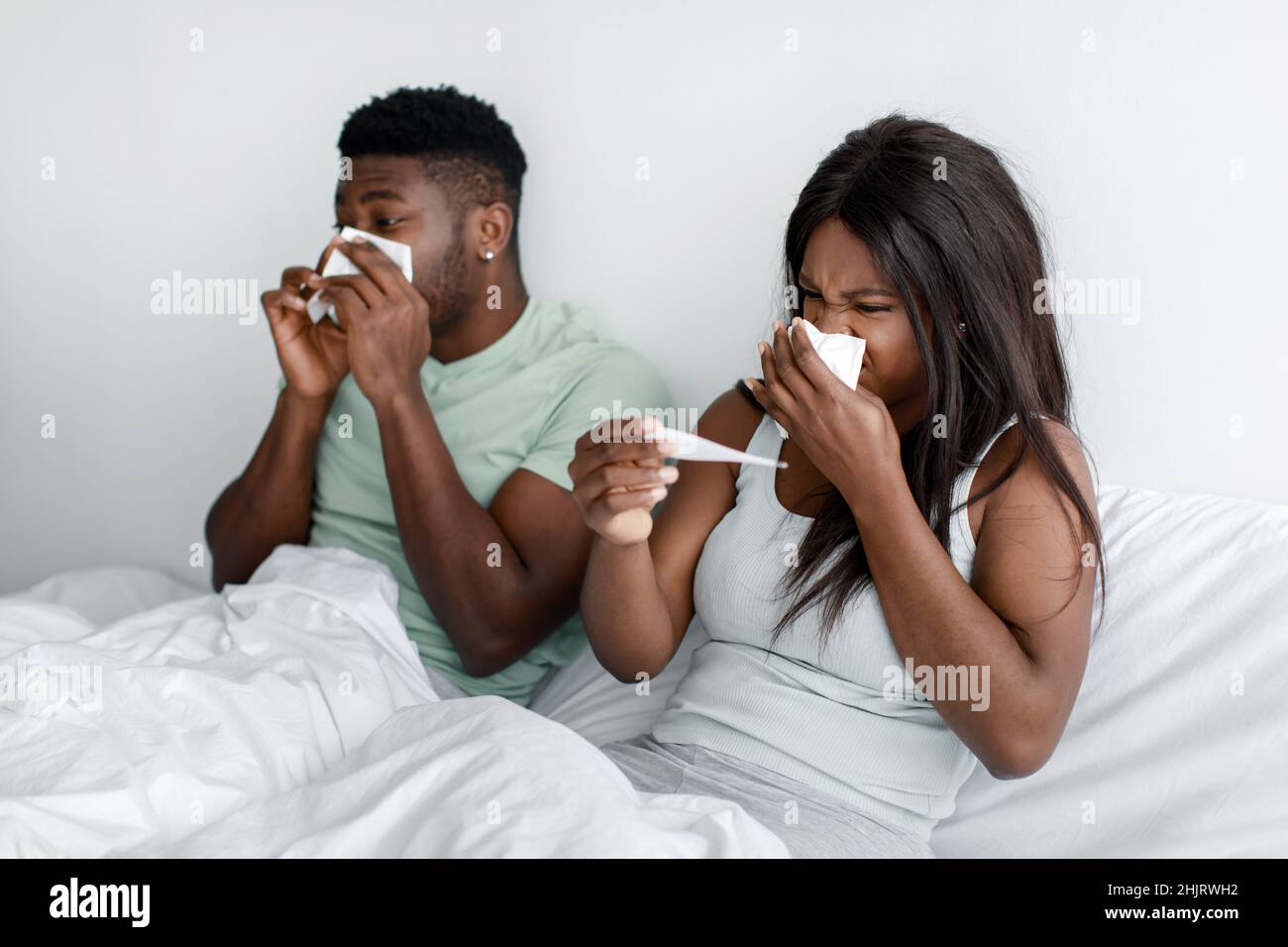 Trauriger junger afroamerikanischer Ehemann und Ehefrau blasen Nase auf Serviette und schauen auf Thermometer auf dem Bett im Schlafzimmer Stockfoto