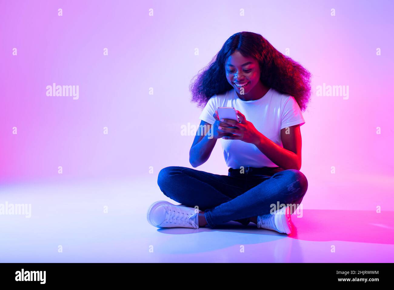 Junge afroamerikanische Dame, die mit gekreuzten Beinen mit dem Handy sitzt, in sozialen Medien surft, mit einem Freund im Neonlicht chattet Stockfoto