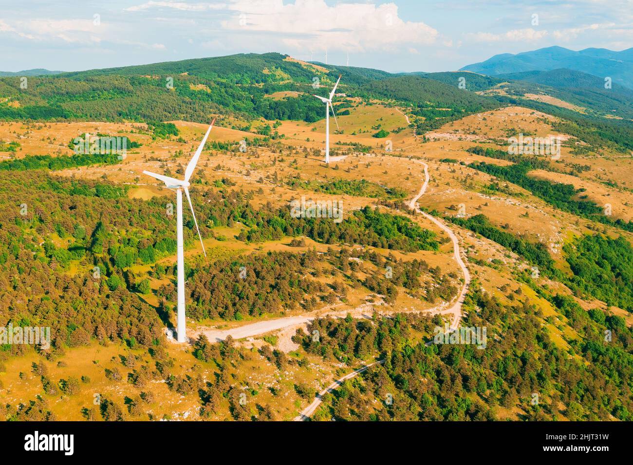 Windmühlen mit großen Klingen werden in Waldbergen betrieben und erzeugen unter bewölktem Himmel am Sommertag saubere Energie von der Drohne aus Stockfoto