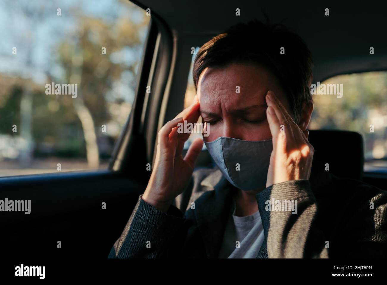 Geschäftsfrau mit starken Kopfschmerzen Schmerzsymptomen und schützender Gesichtsmaske auf dem Rücksitz eines Autos, selektiver Fokus Stockfoto