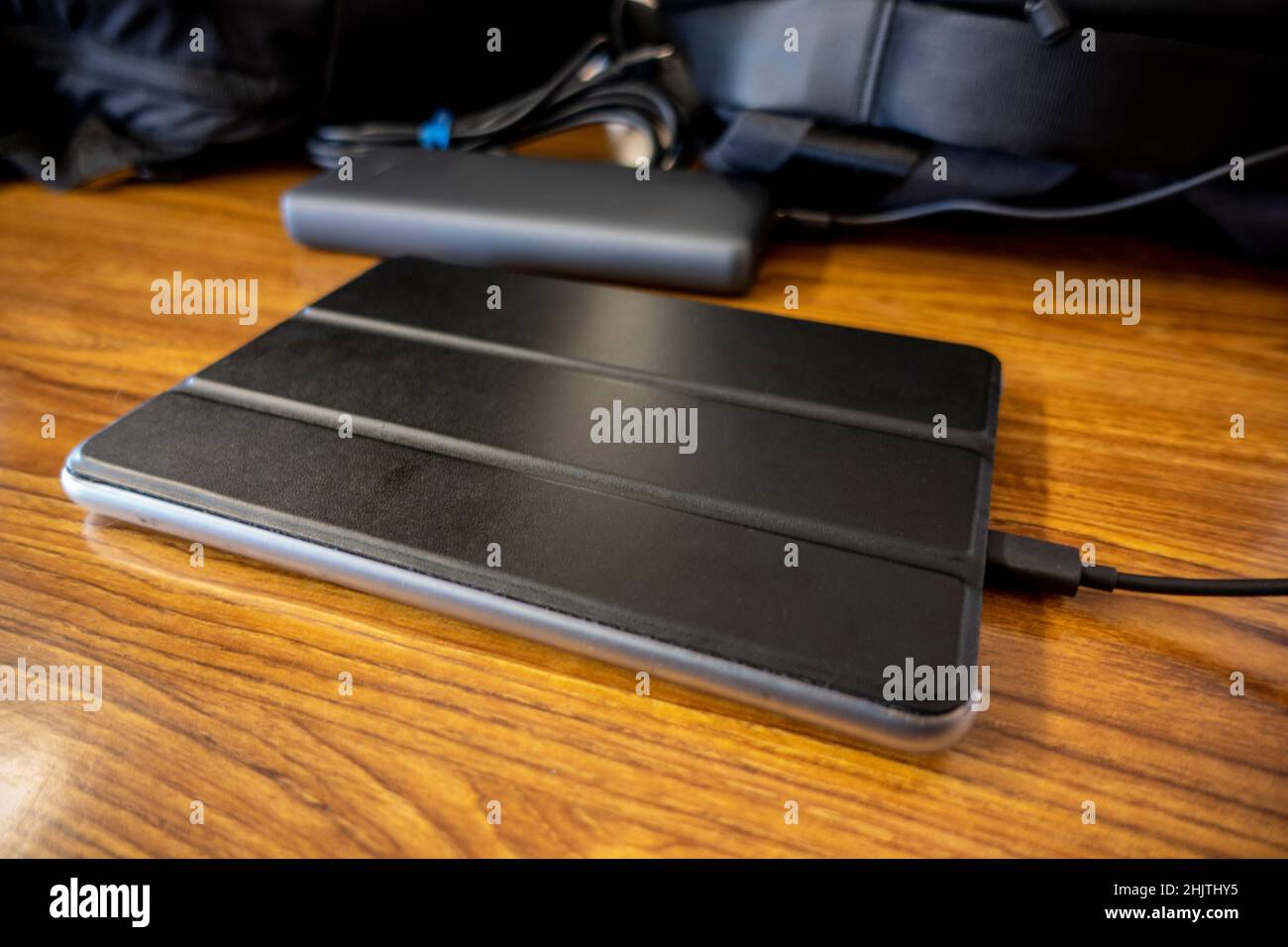 Abgewinkelte Ansicht eines E-Reader-Geräts in einem schwarzen Gehäuse, das auf einem Holztisch aufgeladen wird, mit Rucksäcken im Hintergrund Stockfoto