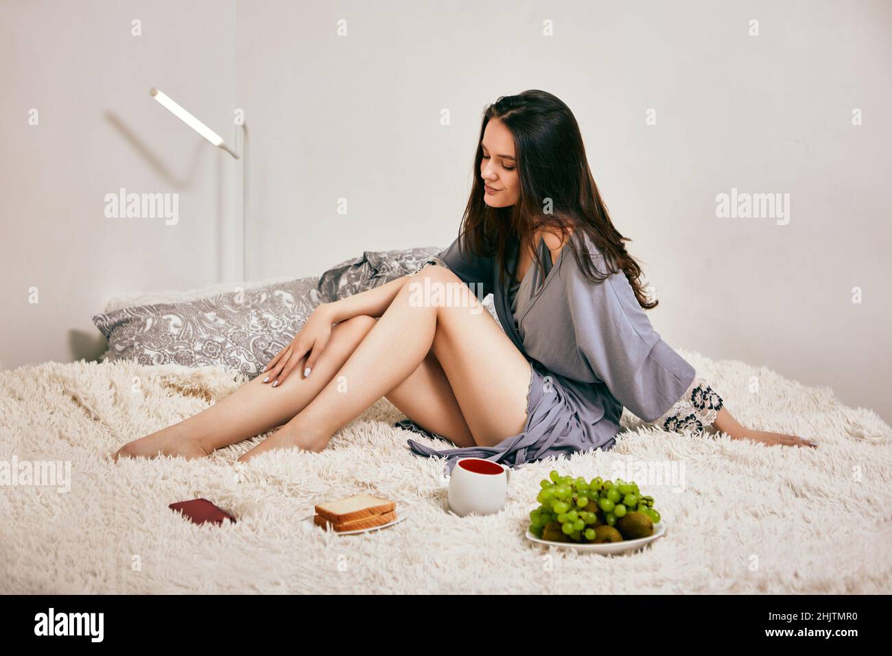 Junge Frau im Morgenmantel mit einer Tasse Kaffee, Obst und einem Sandwich im Bett. Stockfoto