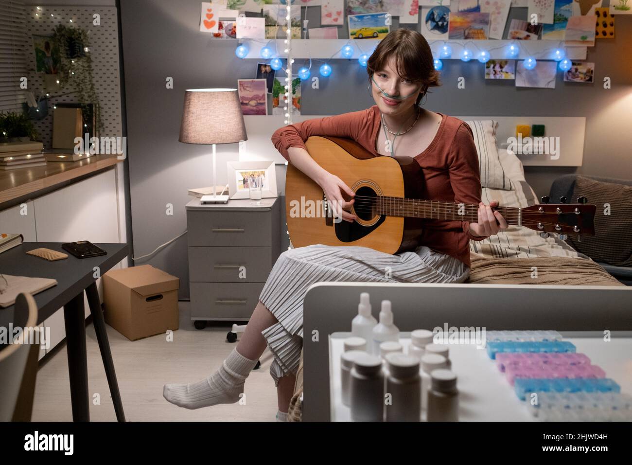 Porträt einer kranken Frau, die während ihrer Musiktherapie im Krankenhaus auf dem Bett sitzt und Gitarre spielt, während sie die Kamera anschaut Stockfoto