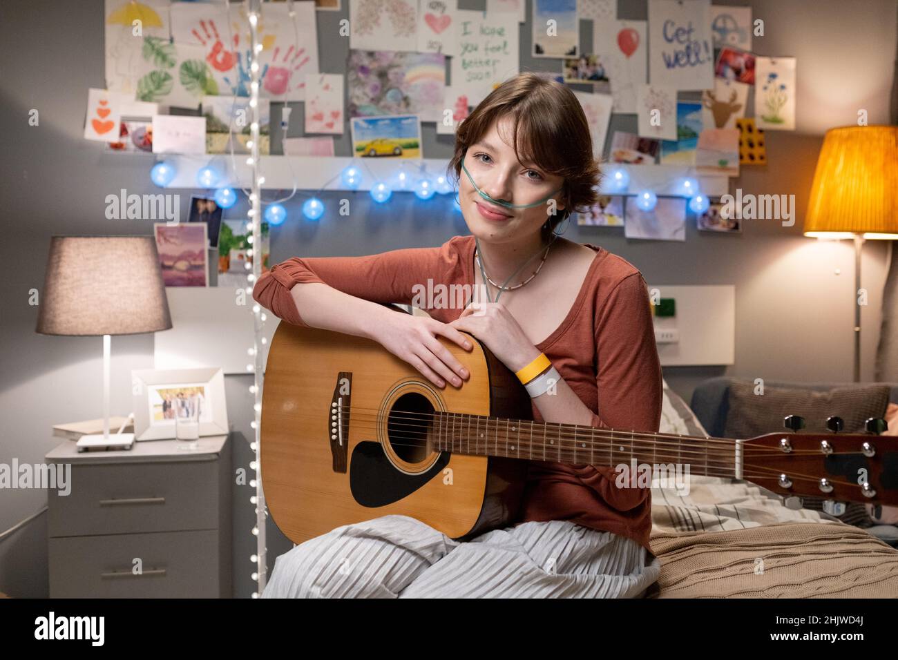 Porträt eines kranken Teenagers, das mit ihrer Gitarre auf dem Bett sitzt und die Kamera anschaut, die sie während ihrer Behandlung genießt, Musikinstrument zu spielen Stockfoto